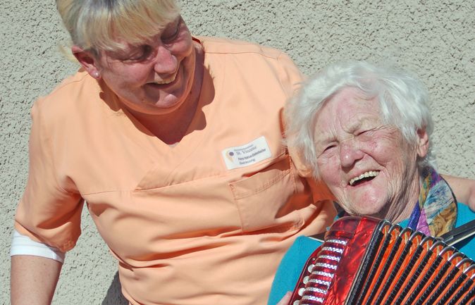 Betreuerin lacht mit Seniorin in Tagesbetreuung beim Akkordeonspielen